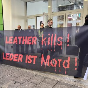 Menschen blockieren den Eingang der Ledermesse in Offenbach. Sie tragen ein Transparent mit der Aufschrift Leather kills bzw. Leder ist Mord. Im Hintergrund ist das Gebäude der Messe Offenbach zu sehen.